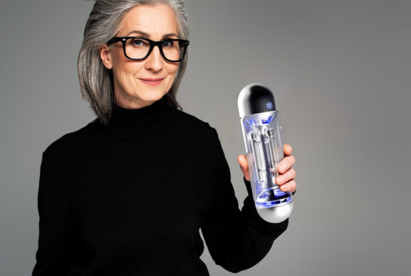 Older woman with glasses holding a modern skincare device, smiling against a neutral background.